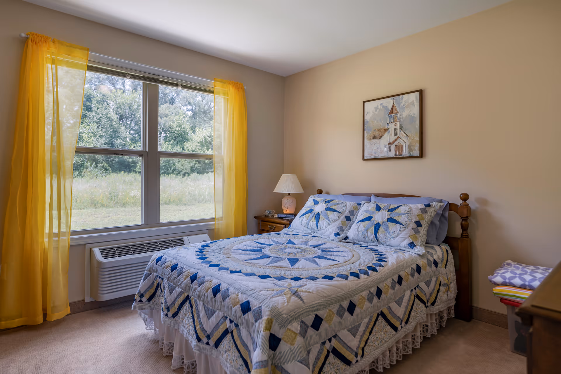 A cozy bedroom with a bed covered in a blue and white patterned quilt and matching pillows. There is a wooden nightstand with a lamp and a small clock next to the bed. A large window with yellow sheer curtains lets in natural light and shows greenery outside. A framed painting of a church hangs on the beige wall above the bed.