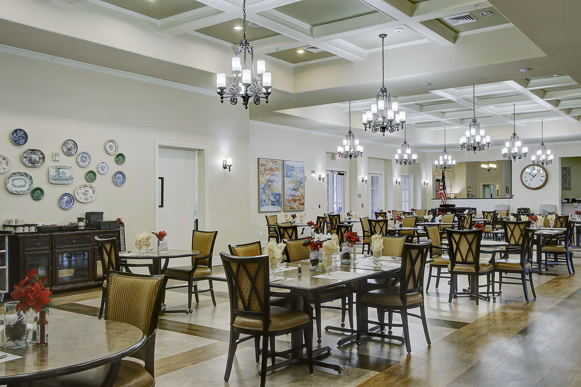 A spacious and well-lit dining room in a senior living facility with multiple tables and chairs arranged neatly. Each table is set with napkins, glasses, and small flower arrangements. The room features elegant chandeliers hanging from a coffered ceiling, decorative plates on one wall, and large windows letting in natural light.