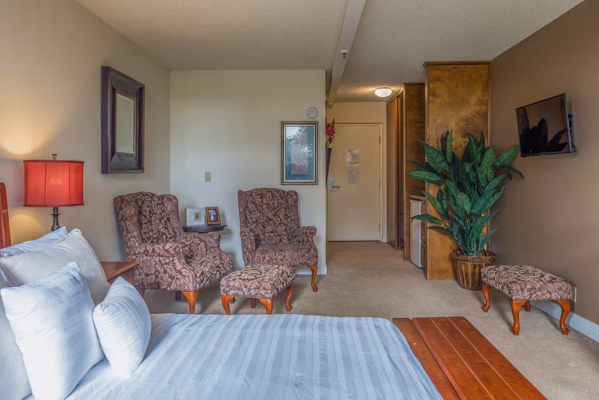 A cozy bedroom in a senior living facility with a neatly made bed featuring white and light blue striped bedding. Two patterned armchairs with a matching ottoman are placed near the wall, accompanied by a small side table with framed photos. A large green potted plant sits near a mounted flat-screen TV on the wall. The room has beige carpeting, a wooden cabinet, and a door leading to another area.
