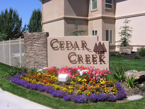 Outdoor view of a landscaped area with a stone and stucco sign that reads 'Cedar Creek' surrounded by colorful flowers and greenery, with part of a building and a white fence in the background.