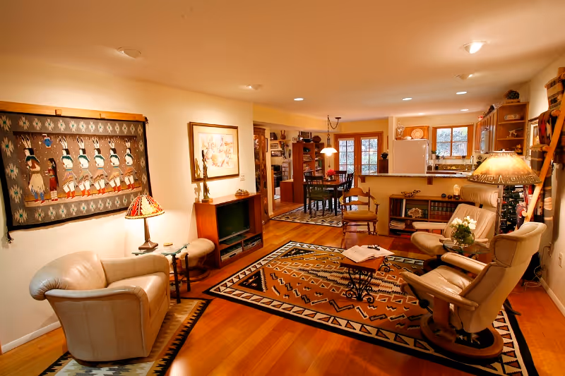 Cozy living room area with wooden floors, leather armchairs, a patterned rug, a small table with an open book, and warm lighting. The room opens into a dining area with a table and chairs, and a kitchen with wooden cabinets and a refrigerator. Decorative wall hangings and lamps add a homey touch.