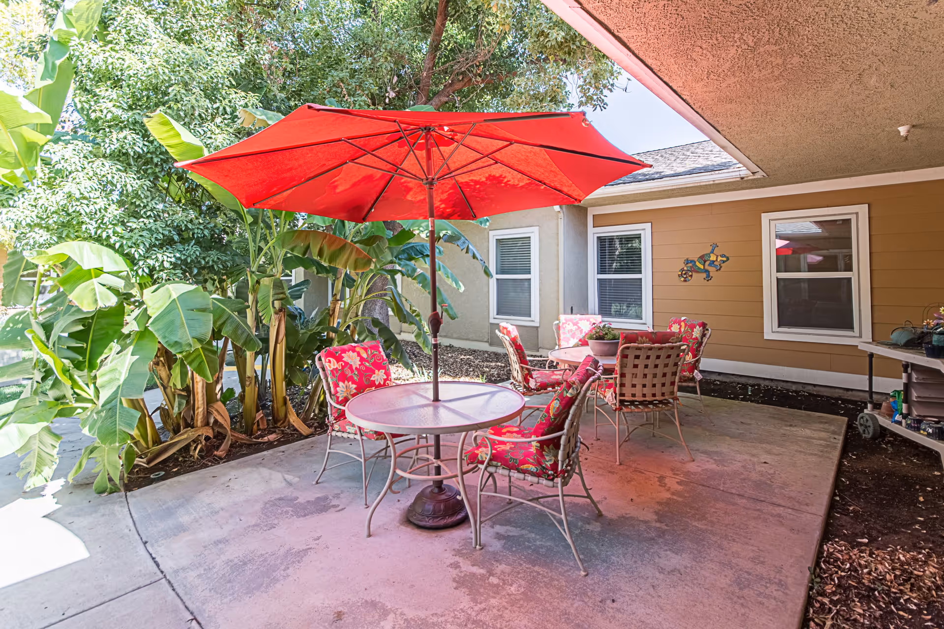 Outdoor patio area with a round table and four chairs featuring red floral cushions, shaded by a large red umbrella. The patio is surrounded by lush green plants and trees, with a beige building wall and windows in the background.