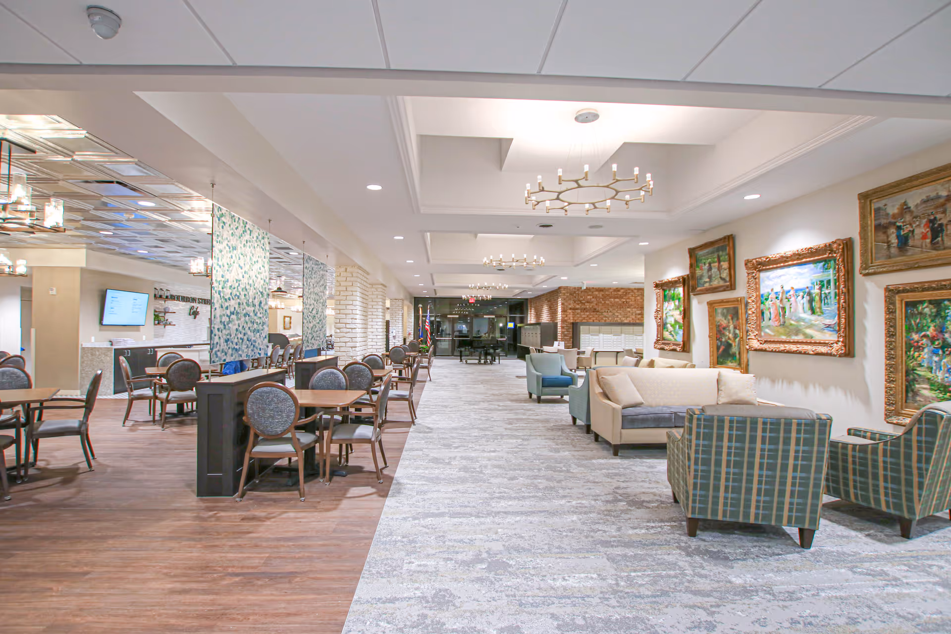Spacious interior of a senior living facility featuring a dining area with multiple tables and chairs on the left, and a comfortable seating area with sofas and armchairs on the right. The walls are decorated with framed paintings, and modern chandeliers hang from the ceiling. The floor transitions from wood in the dining area to carpet in the seating area.