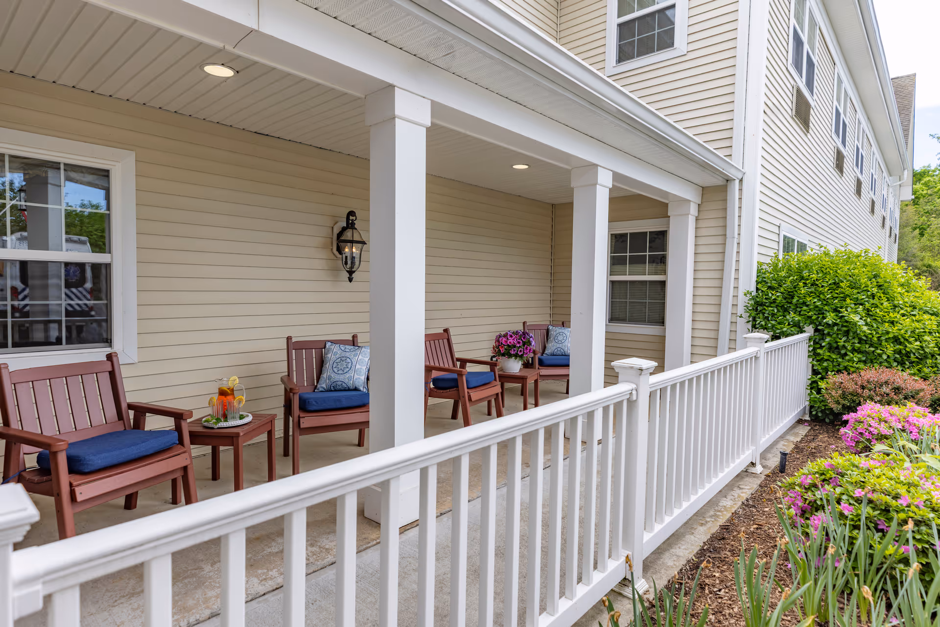 Covered outdoor porch area with wooden chairs featuring blue cushions and decorative pillows, small tables with a pitcher of iced tea and a potted plant, white railing, beige siding on the building, and colorful flowers and green bushes along the garden bed.