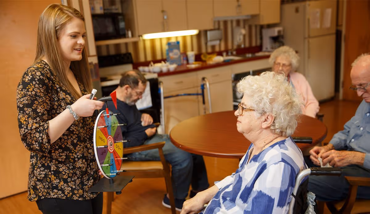 A young woman holding a colorful spinning wheel is engaging with elderly residents seated around a table in a room with a kitchen area in the background. The elderly individuals appear to be participating in an activity or game.
