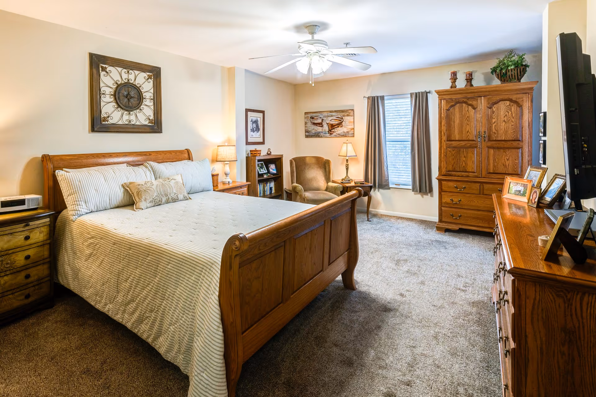 Well-lit bedroom with a wooden bed, matching dressers and armoire, an armchair, ceiling fan, and a window with curtains.