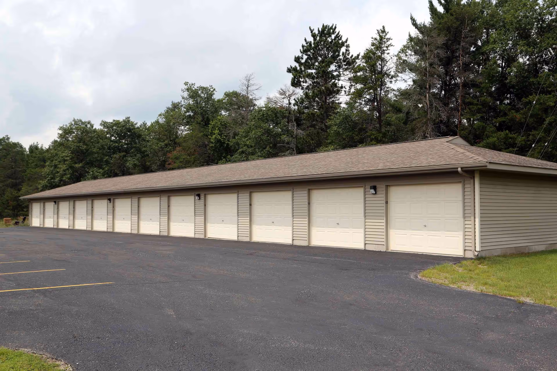 A long building with multiple beige garage doors numbered 3 to 12, situated next to a paved parking area with yellow lines. The building is surrounded by green trees and grass under a cloudy sky.