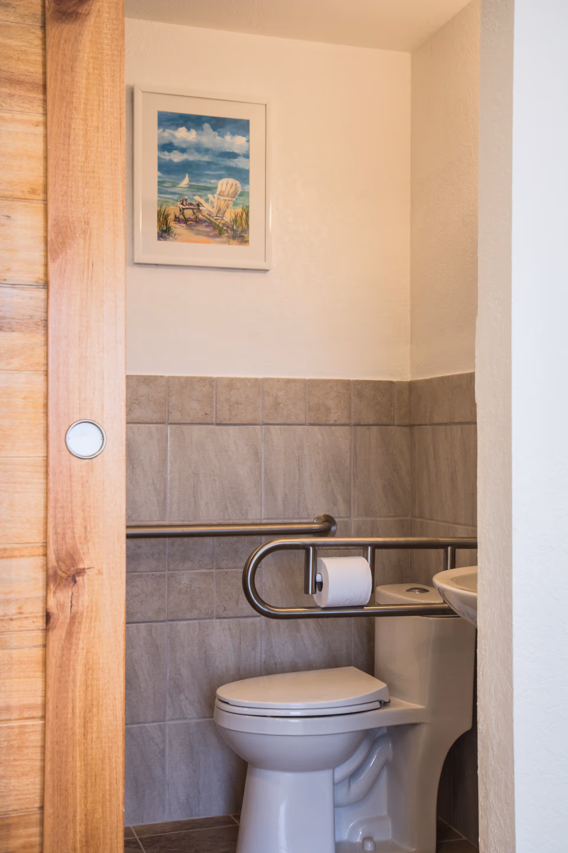 Accessible bathroom with a toilet, metal grab bars (one holding a roll of toilet paper), a sink, tiled walls, and a framed beach painting.