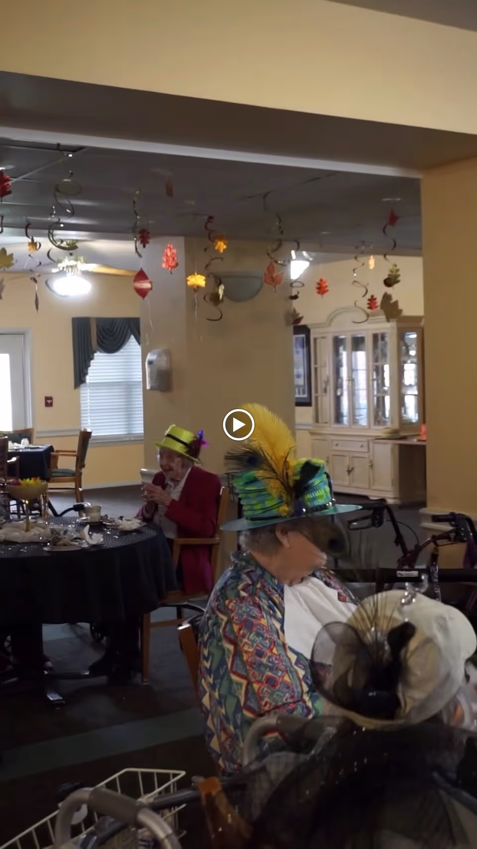 Two elderly women wearing colorful festive hats seated at a table in a decorated dining area with hanging autumn leaf decorations and a china cabinet in the background.