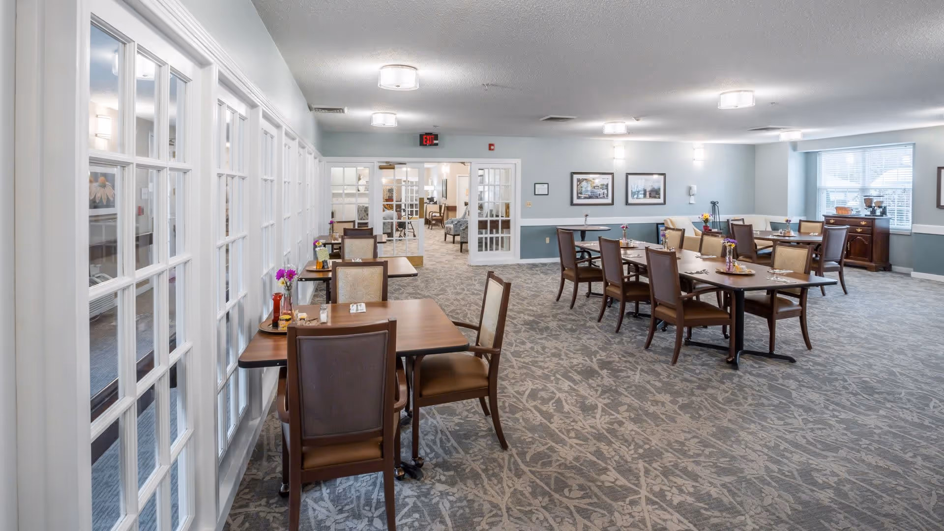 A spacious dining room with multiple wooden tables and chairs arranged neatly. The room has large windows letting in natural light, light blue walls with white trim, framed pictures on the walls, and a carpeted floor with a subtle pattern. There are glass-paneled double doors leading to another room in the background.
