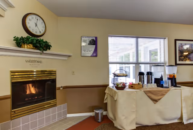 Community living room with a lit fireplace, wall clock, and a table set up with coffee and refreshments by a window.