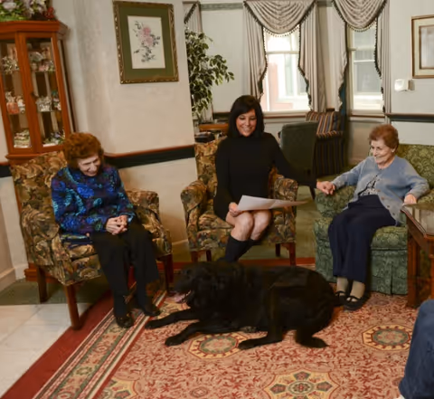 Three women sitting in a cozy living room with floral and patterned armchairs. Two elderly women are seated on either side of a younger woman who is holding a piece of paper. A large black dog is lying on a patterned rug in front of them. The room has framed artwork on the walls, a glass cabinet with decorative items, and windows with draped curtains.
