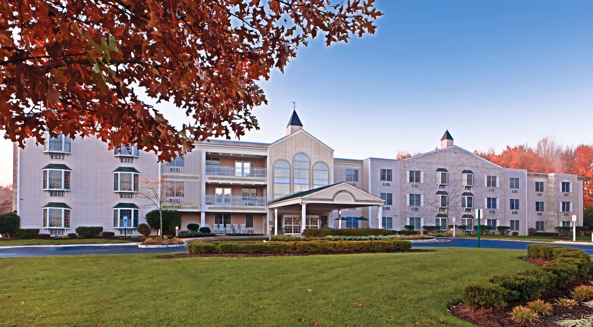 Front exterior of a multi-story senior living facility with a covered entrance, balconies, and landscaped lawn in autumn.