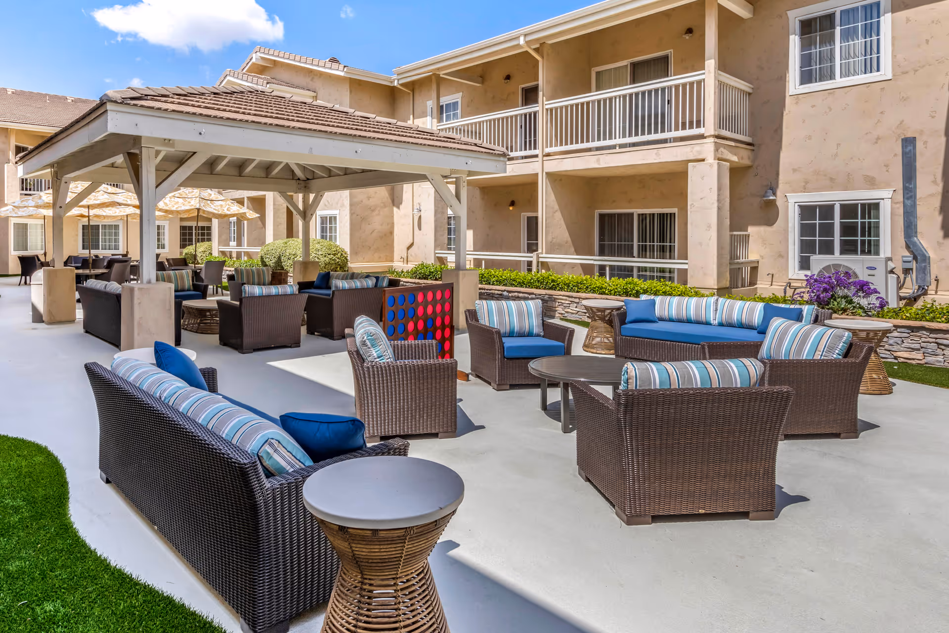 Outdoor patio area at Meadowbrook of Agoura Hills featuring multiple wicker seating arrangements with blue and striped cushions, a covered gazebo with additional seating, a large Connect Four game, and a beige multi-story building in the background under a blue sky with some clouds.
