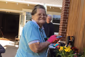 An elderly woman wearing a light blue shirt and pink gloves is gardening outdoors near a wooden fence with yellow flowers in front of her. An elderly man stands in the background near a brick wall and an open doorway.
