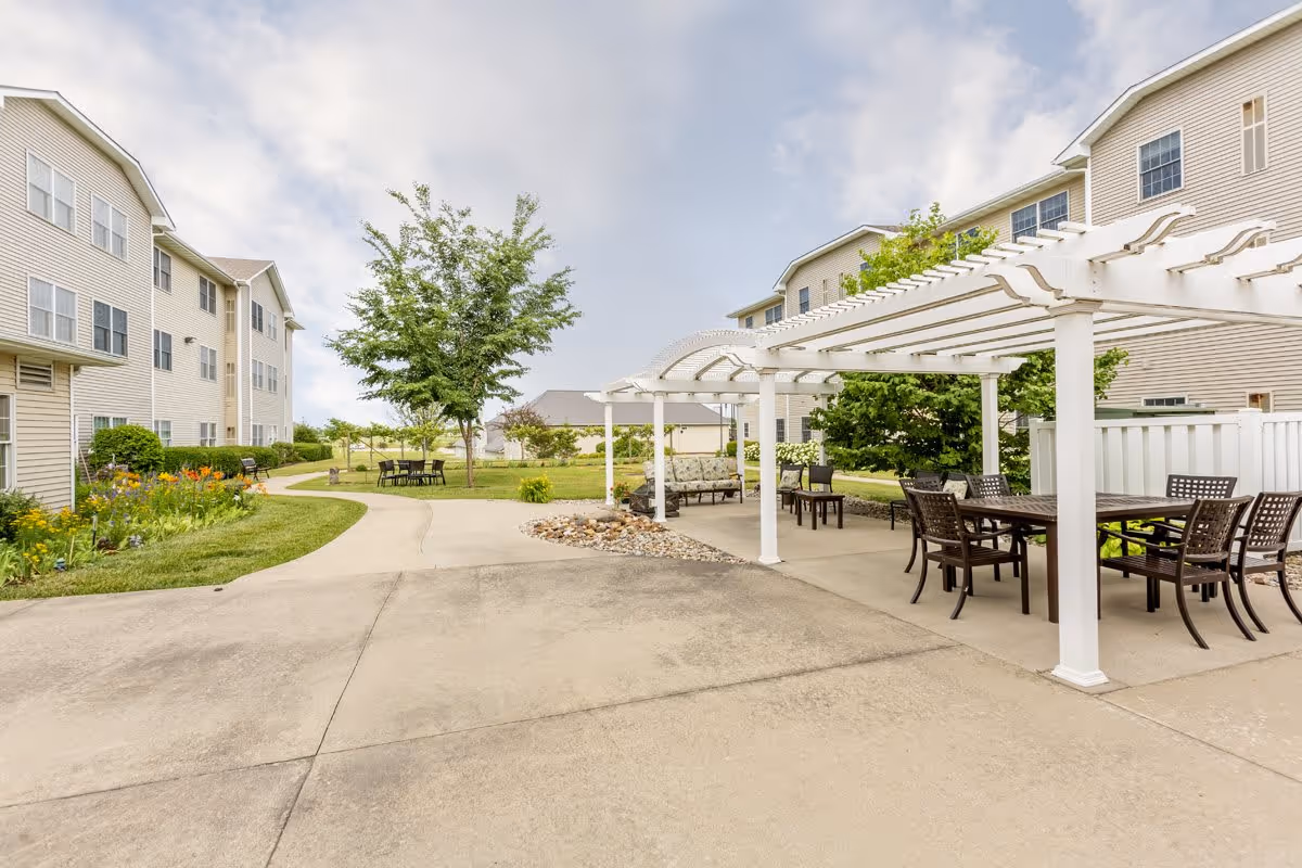Outdoor patio area at a senior living facility with a white pergola covering several tables and chairs. There are two multi-story beige buildings on either side, a concrete walkway, green grass, trees, and flower beds under a partly cloudy sky.