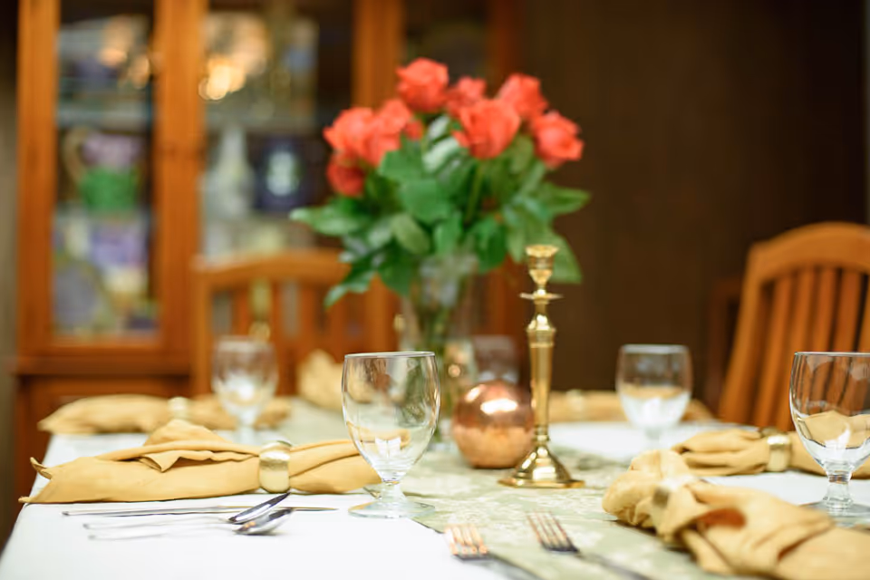 A dining table set with clear water glasses, beige cloth napkins with gold napkin rings, silverware, a gold candlestick, and a vase of red roses in the background. Wooden chairs and a glass-front cabinet are visible behind the table.