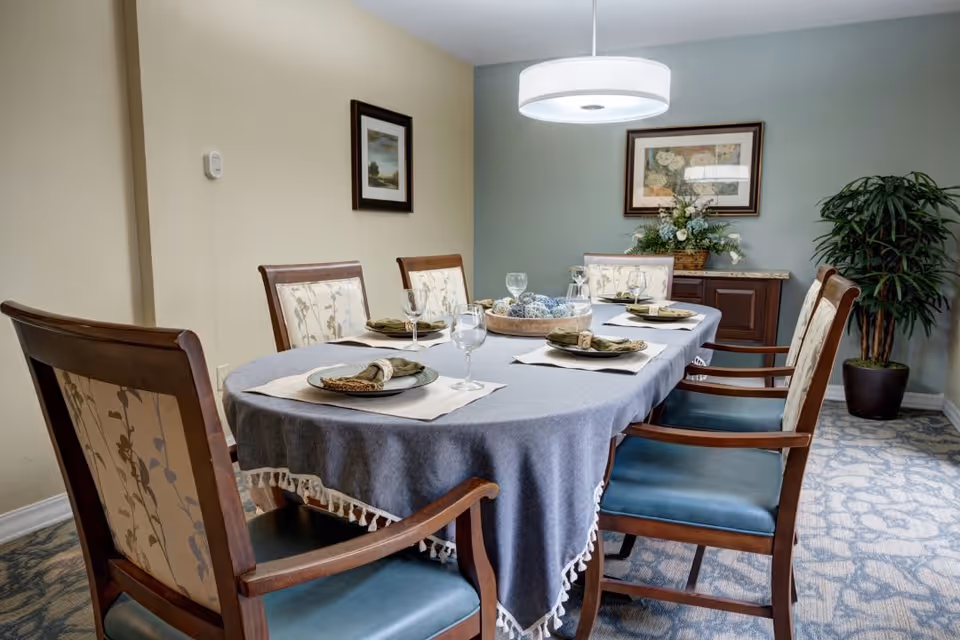 A dining room with a rectangular table covered with a blue tablecloth and set with plates, napkins, and glasses. Six wooden chairs with cushioned seats and backs surround the table. The room has light-colored walls, a framed picture on each wall, a sideboard with a floral arrangement, a potted plant, and a modern circular ceiling light fixture.