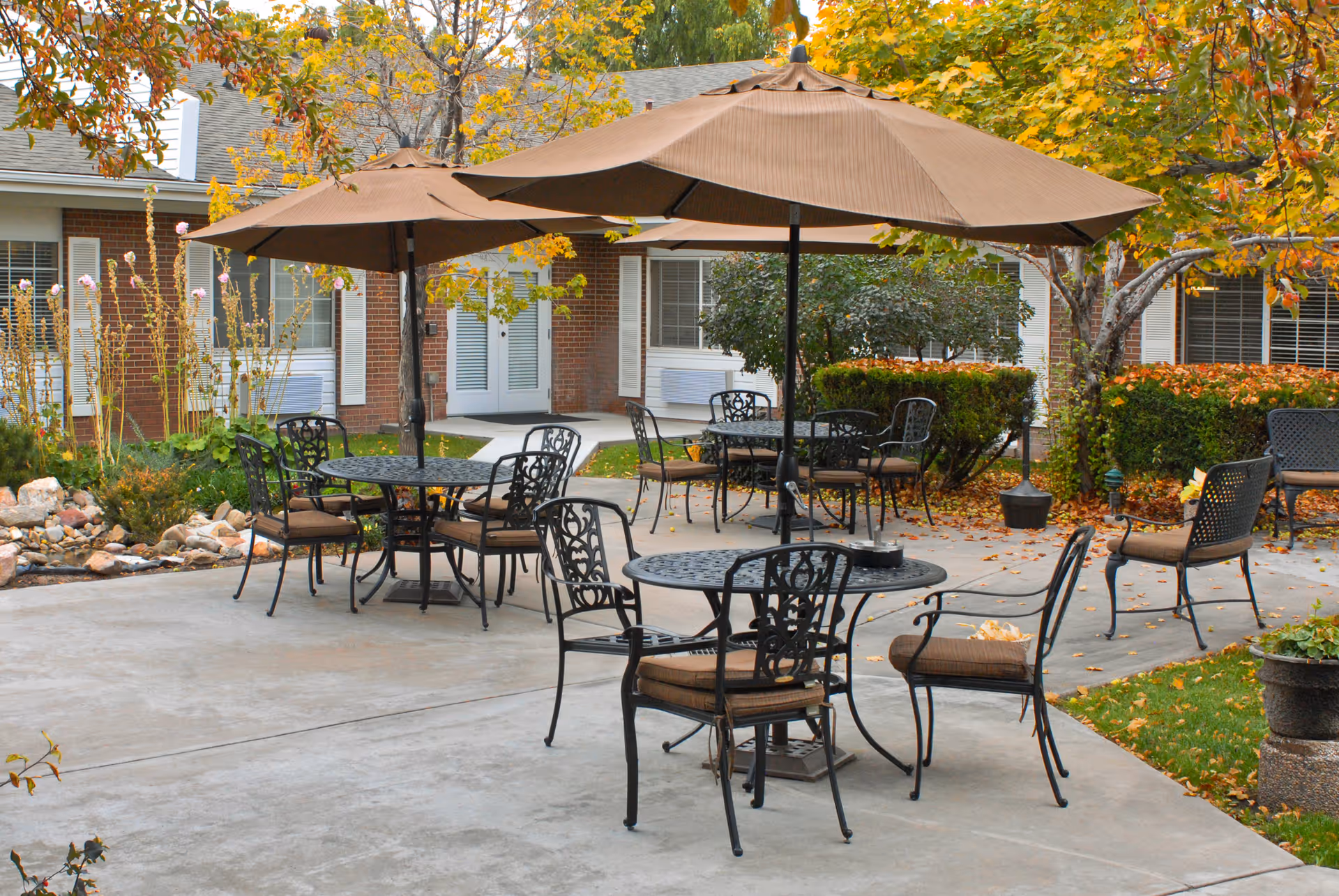 Outdoor patio area with several round metal tables and cushioned chairs under large brown umbrellas. The patio is surrounded by greenery, bushes, and trees with autumn leaves. A brick building with windows and doors is visible in the background.