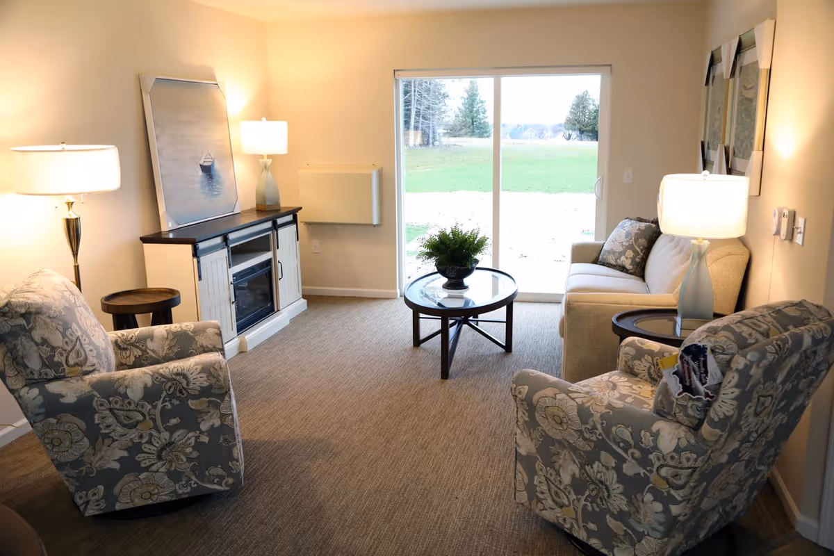 A cozy living room with two patterned armchairs, a beige sofa, a round glass coffee table with a plant centerpiece, two side tables each with a lamp, a cabinet with a painting and lamp on top, and a sliding glass door showing a green outdoor area.