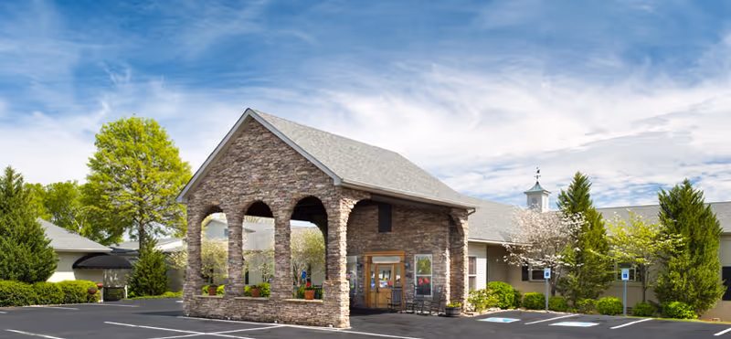 Front entrance of a stone-clad building with an arched porte-cochere, parking spaces, and landscaped trees under a blue sky.