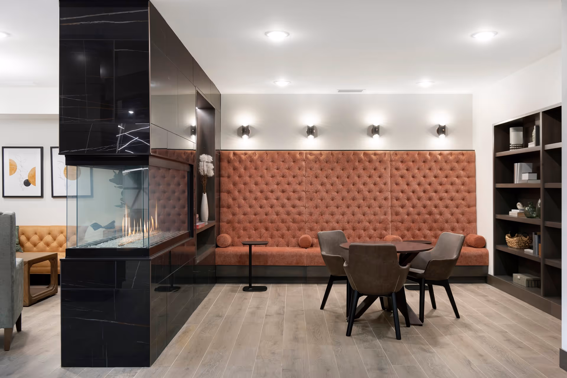 Modern lounge area with a long tufted orange-brown bench along the wall, a round table with four brown chairs, a black marble fireplace with glass panels, and a bookshelf with decorative items. The room has light wood flooring and white walls with wall-mounted lights.