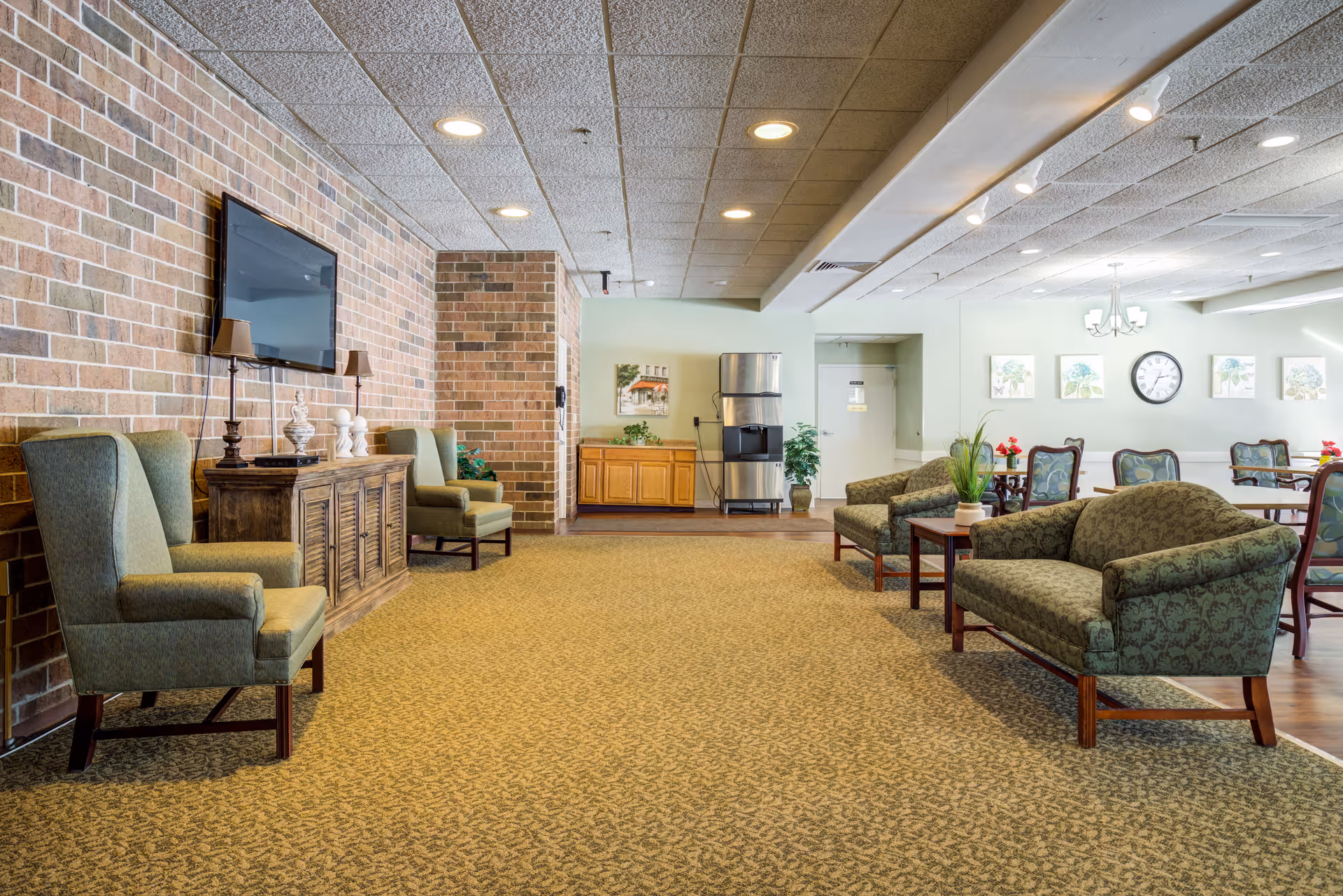 Spacious senior living lounge with upholstered chairs and sofas, a wall-mounted TV, and a dining area in the background.