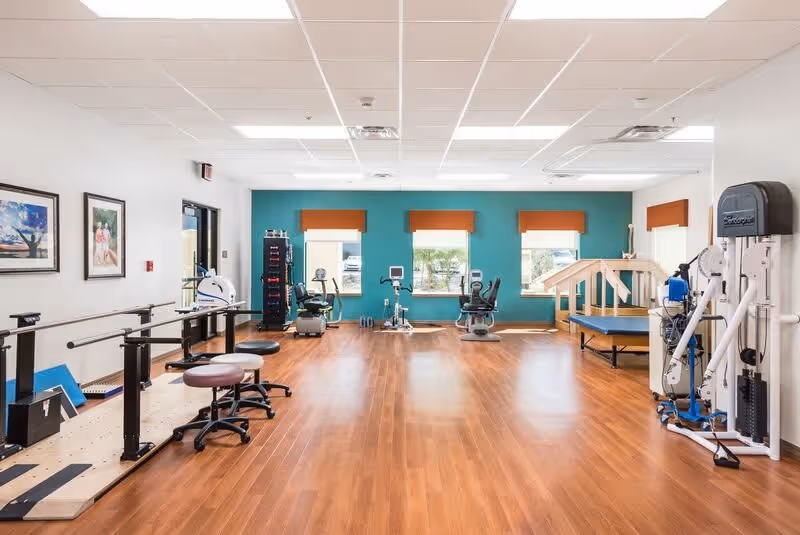 A bright rehabilitation therapy room with parallel bars, exercise bikes, strength equipment, and wood floors.