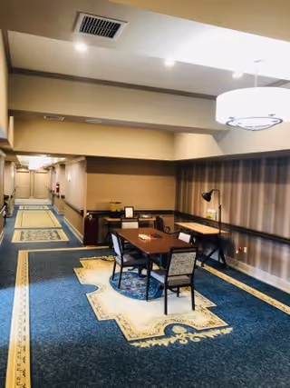 A well-lit hallway in an assisted living facility with a blue and beige patterned carpet. In the foreground, there is a small seating area with a table and four chairs, a desk with a lamp, and a large ceiling light fixture. The hallway extends into the background with doors and wall-mounted lights along the sides.