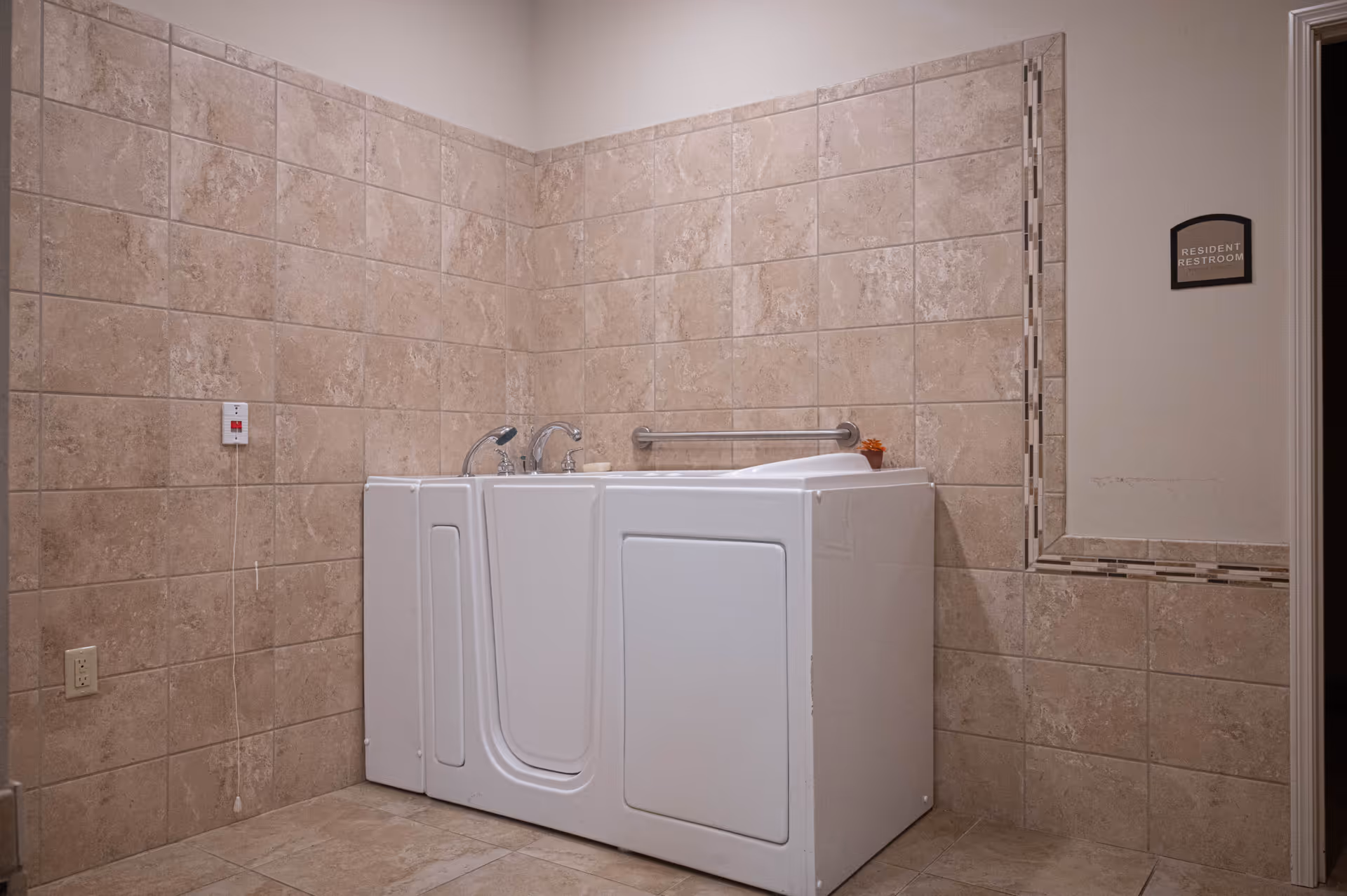 A walk-in bathtub with a door and faucet installed in a tiled bathroom corner. The walls and floor are covered with beige tiles. A small sign on the wall near the doorway reads 'Resident Restroom.'