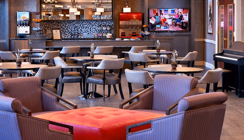 A cozy common area in an assisted living facility featuring several round tables with chairs, a red cushioned ottoman surrounded by striped armchairs, a beverage station with a soda fountain and juice dispenser, a wall-mounted television showing a talk show, and a piano in the corner.