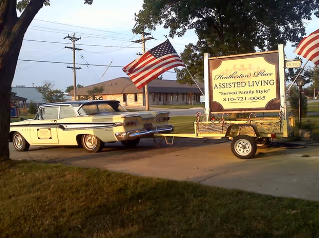A vintage cream-colored car is parked on a driveway next to a trailer carrying a large sign for Heatherton Place Assisted Living, which includes the tagline 'Served Family Style' and a phone number. Two American flags are displayed, one attached to the car and one near the sign. Trees and buildings are visible in the background.