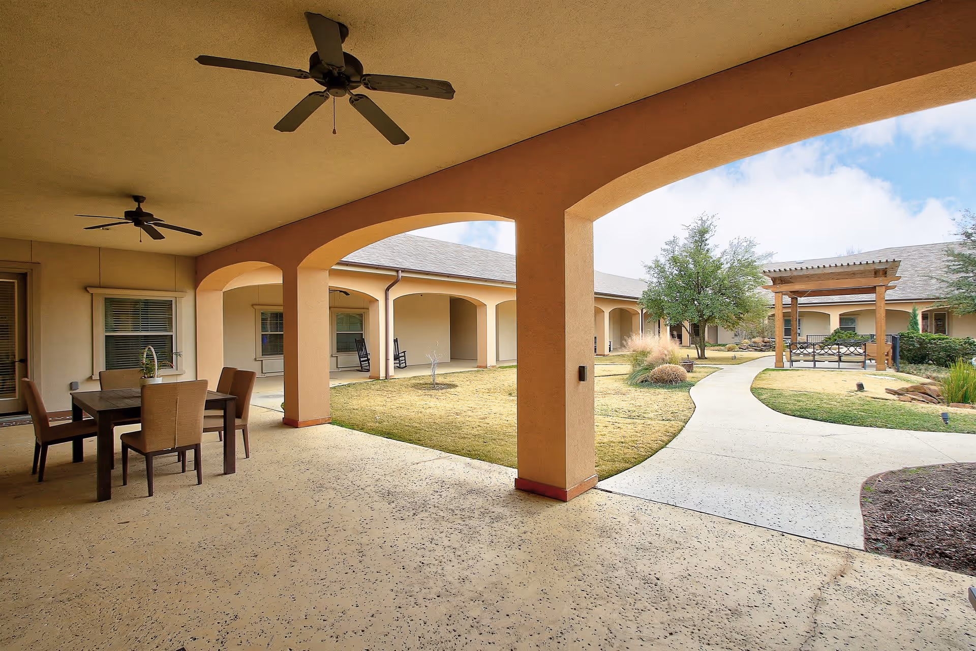 Covered outdoor patio area with ceiling fans and a dining table with chairs, overlooking a landscaped courtyard with a walking path, trees, and a wooden pergola bench at Volante Senior Living of Grapevine.