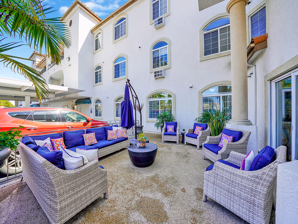 Outdoor seating area at San Clemente Villas featuring wicker furniture with blue cushions and colorful pillows arranged around a circular black table. The area is adjacent to a white multi-story building with several windows and a column. A red car is parked nearby under a covered parking area, and there are some green plants and a palm tree visible.