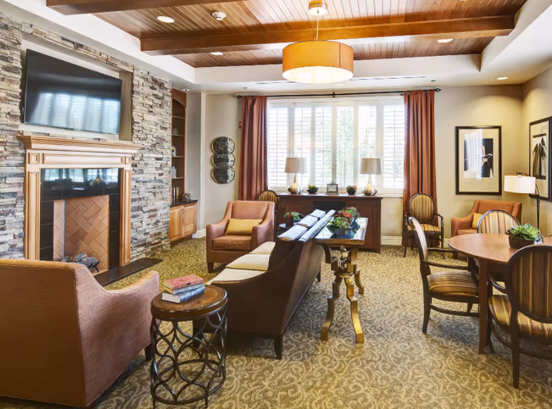 Senior living lounge with stone fireplace, TV, and multiple seating areas by large windows.