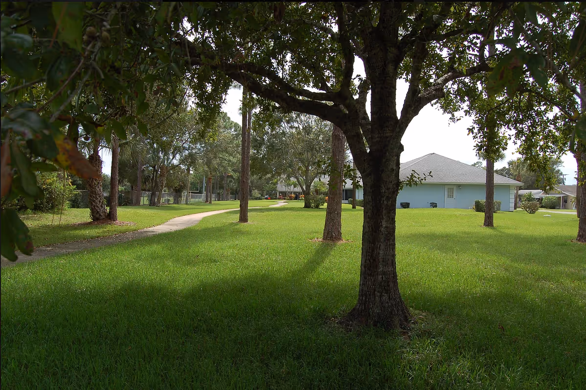 Grassy lawn with trees and a paved path leading toward single-story buildings under a shaded canopy.