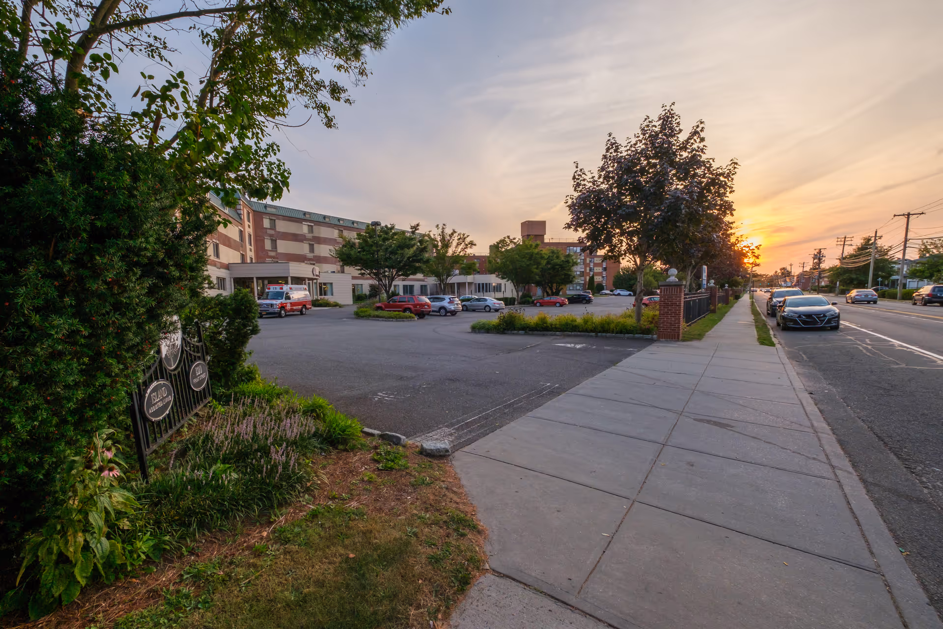 Sidewalk and parking lot leading to the front of a multi-story assisted living building at sunset.
