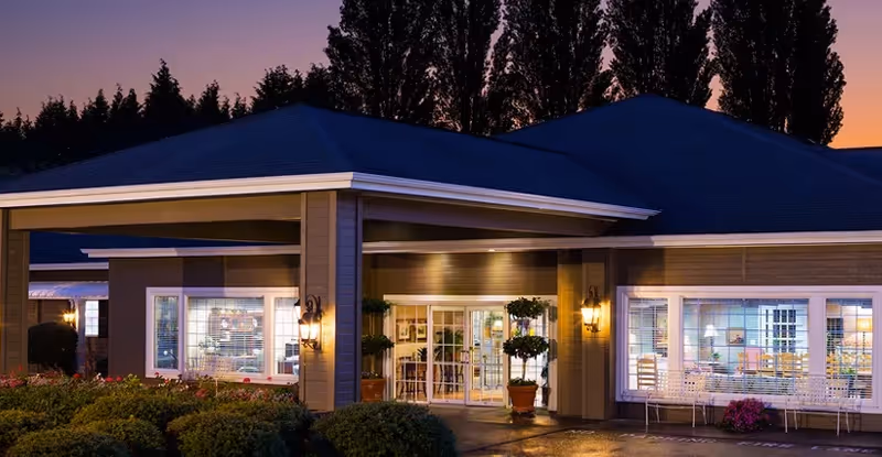 Exterior view of Canterbury Gardens Memory Care Community building at dusk with warm lights glowing from inside, surrounded by bushes and potted plants near the entrance.