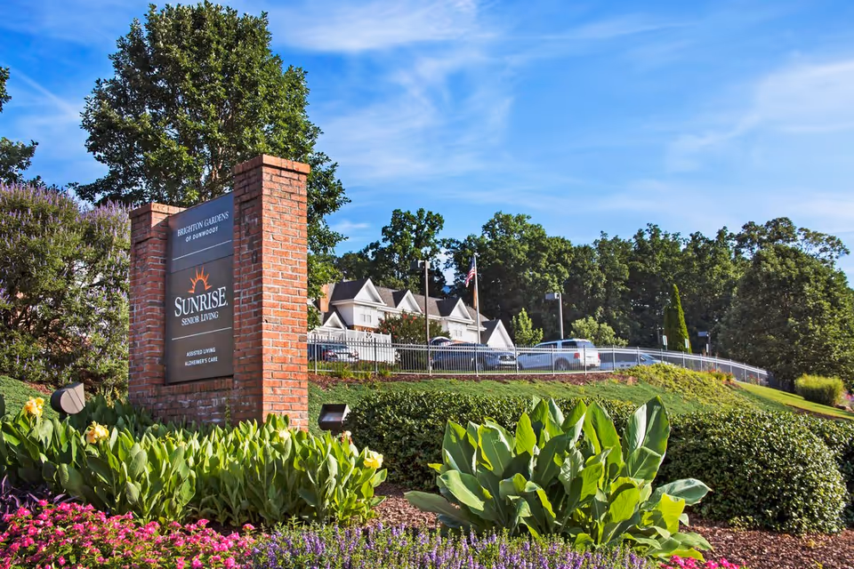 Outdoor view of Brighton Gardens of Dunwoody senior living facility sign made of brick with landscaping including green plants and colorful flowers in front, with a building, parked cars, trees, and a blue sky in the background.