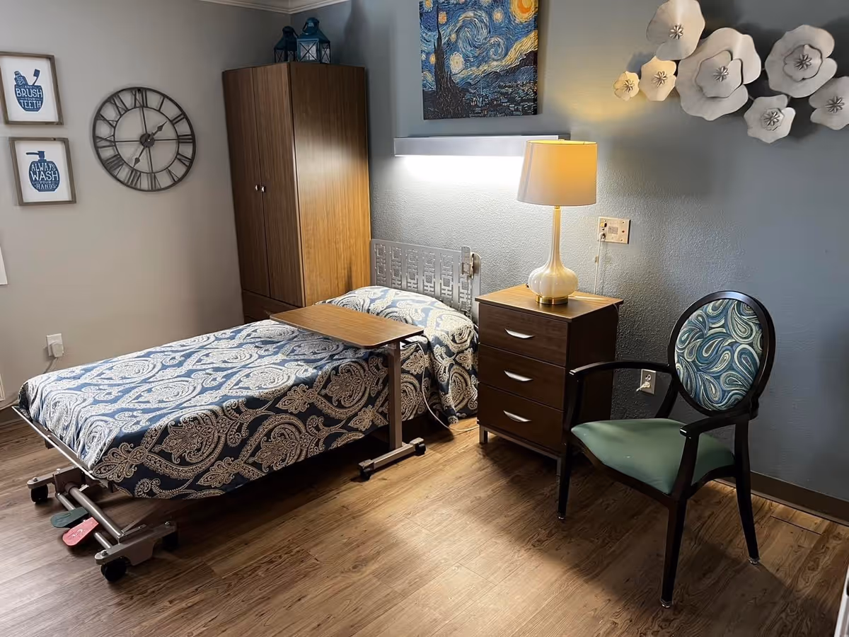 A neatly arranged bedroom in a skilled nursing facility featuring a single hospital-style bed with a blue and white patterned bedspread. Next to the bed is a wooden nightstand with a lamp, and a decorative chair with a patterned cushion. The room has wooden flooring, a wooden wardrobe, a large wall clock, framed wall art with hygiene reminders, and decorative white flower wall hangings.