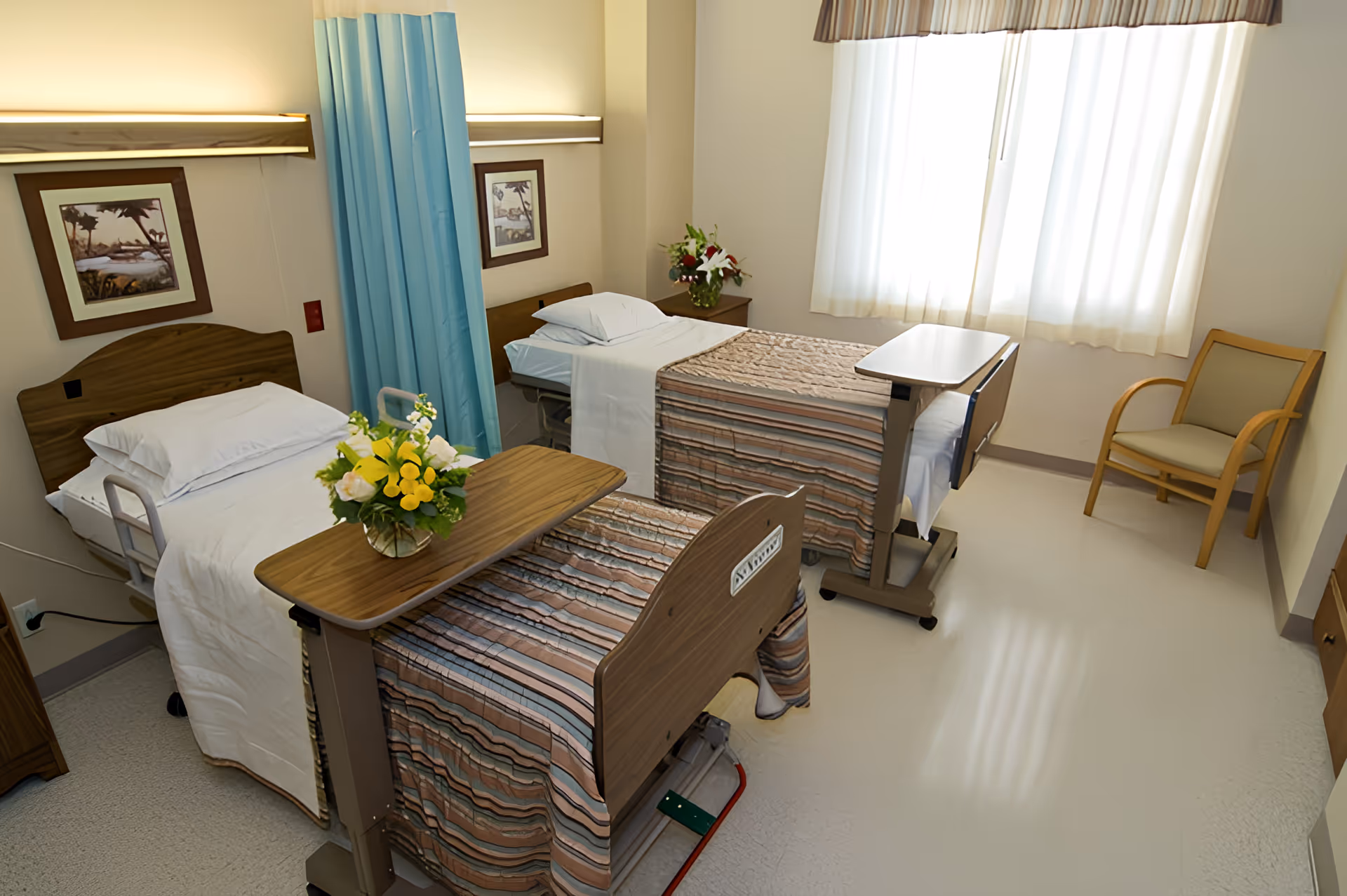 A clean and well-lit room with two hospital-style beds separated by a blue privacy curtain. Each bed has white sheets, pillows, and a striped bedspread. There are two wooden overbed tables, one with a vase of yellow and white flowers. The room has a window with sheer white curtains, a wooden chair, and framed artwork on the walls.