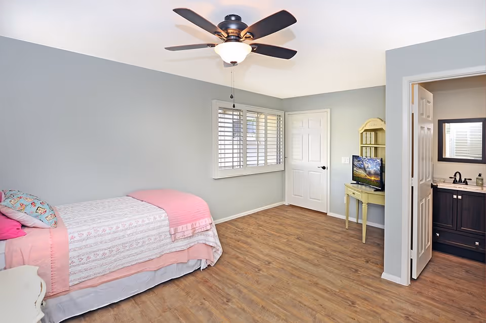 Bright bedroom with a single bed, ceiling fan, small yellow desk with a TV, wood-look floors, and an open doorway to a bathroom.
