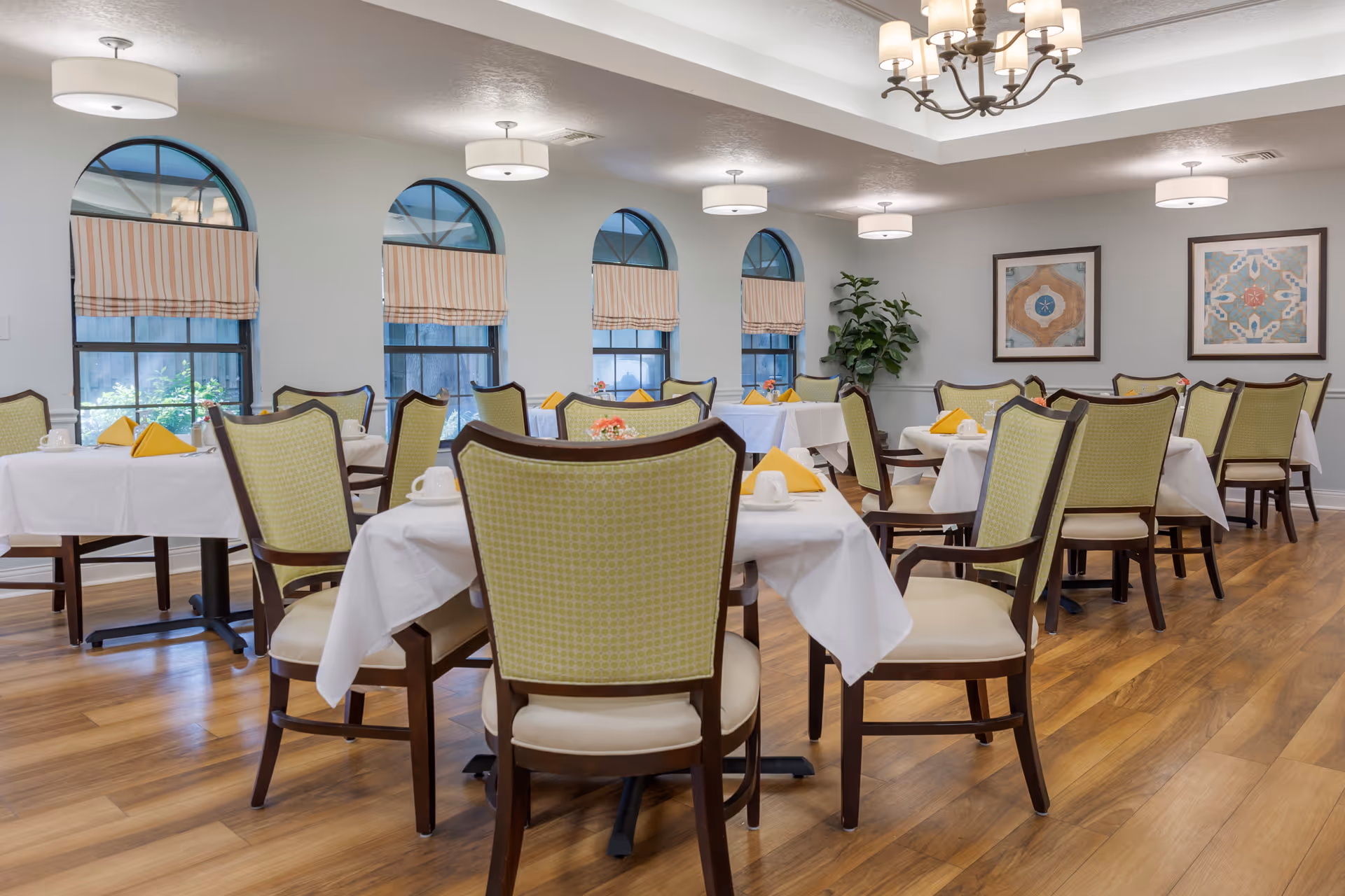 Well-lit dining room with tables set with white linens and yellow napkins, green-upholstered chairs, arched windows and hardwood floors.