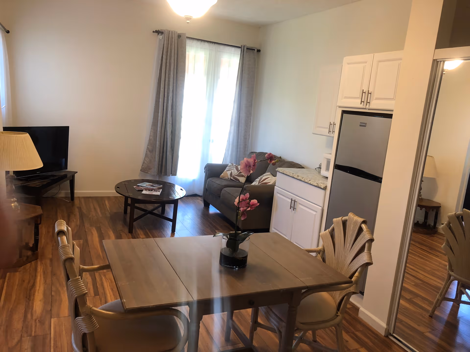 Interior view of a senior living facility room with a wooden dining table and four chairs in the foreground, a small gray couch with patterned pillows near a window with gray curtains, a round wooden coffee table with magazines, a flat-screen TV on a stand, a lamp on a side table, and a kitchenette area with white cabinets and a small refrigerator.