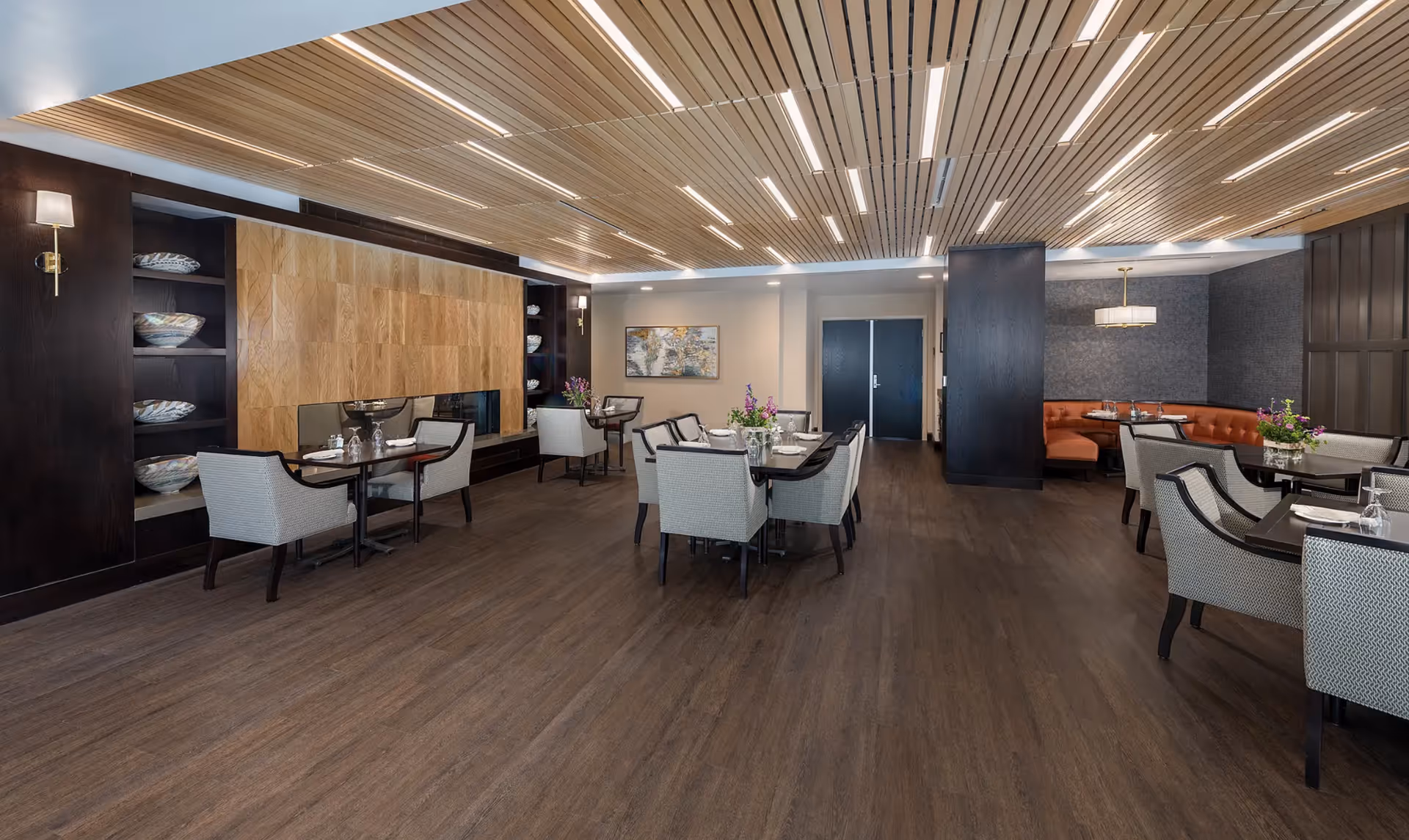 A modern dining room with multiple tables and chairs arranged neatly. The room features a wooden floor, a decorative wooden ceiling with linear lighting, and a wall with built-in shelves holding decorative bowls. There is a large wooden panel on one wall, a painting, and a cozy seating area with an orange cushioned bench and a hanging light fixture.
