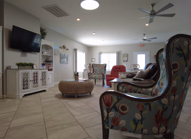 Bright senior living lounge with patterned armchairs, a wicker coffee table, TV and additional seating on a tiled floor.