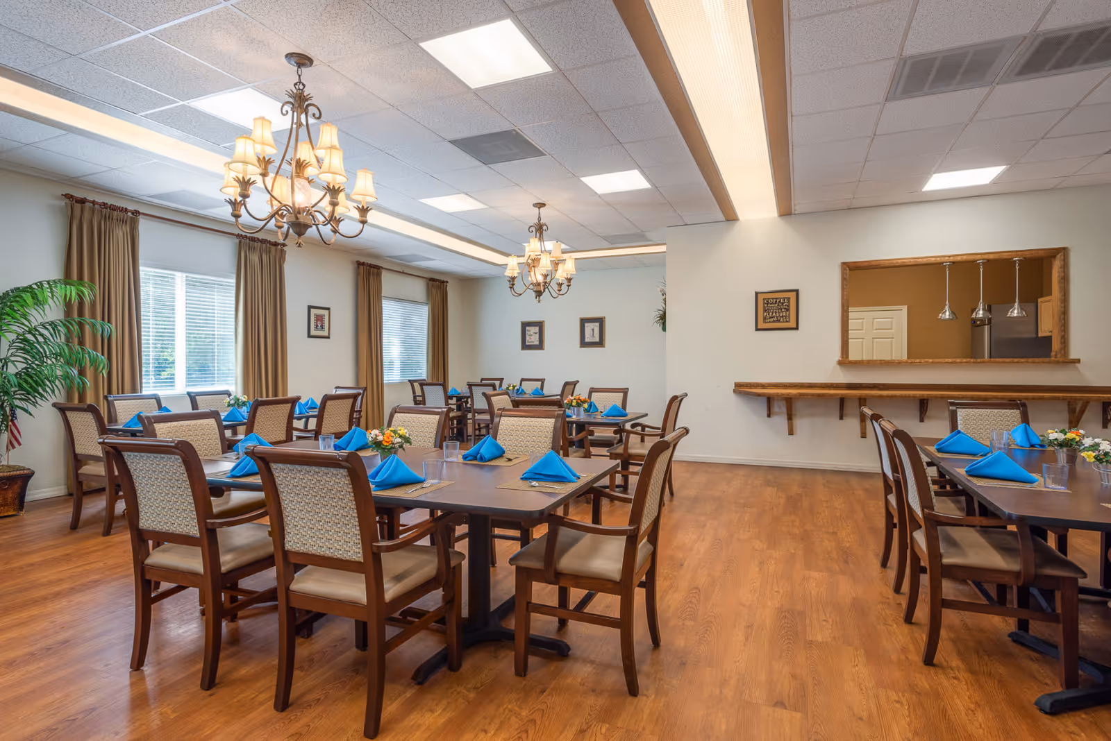 A dining room in a senior living facility with several wooden tables and chairs arranged neatly. Each table is set with blue folded napkins, placemats, and small flower arrangements. The room has large windows with brown curtains, two ornate chandeliers hanging from the ceiling, and a wooden floor. There is a pass-through window with pendant lights on the right wall.