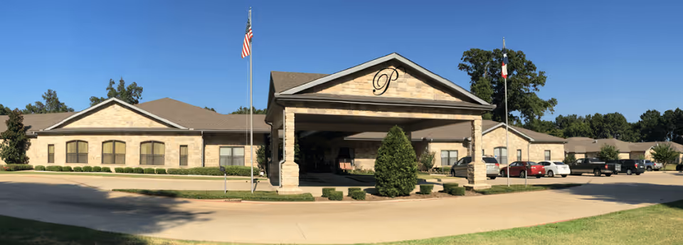 Front exterior view of Prestige Estates Assisted Living & Memory Care building with a covered entrance, stone facade, multiple windows, two flagpoles with American and Texas flags, and a parking area with several cars.