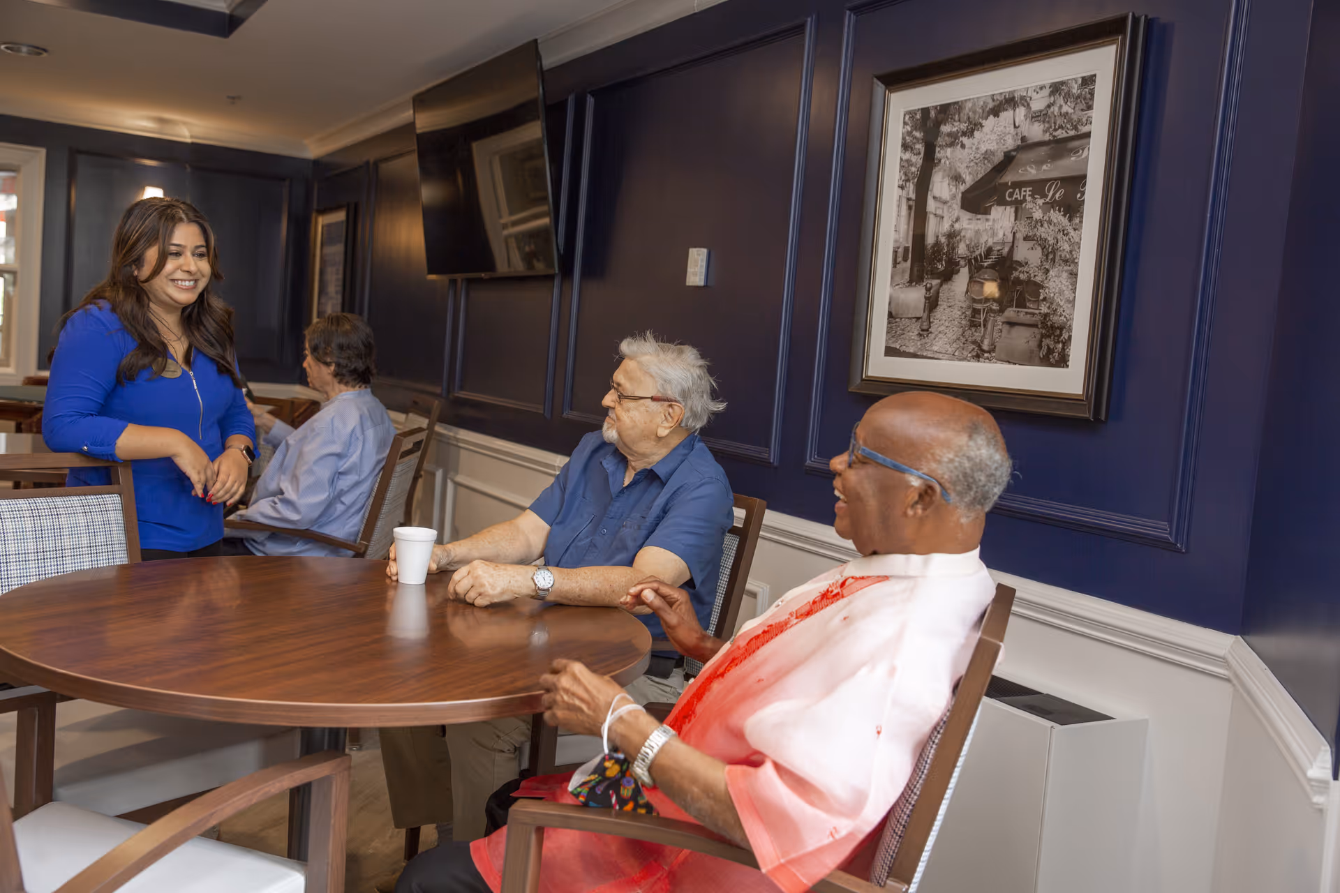 A group of three elderly people sitting around a round wooden table in a room with dark blue walls and white trim. A woman in a blue top is standing and smiling while interacting with the seated individuals. There is a framed black and white picture on the wall and a flat-screen TV mounted above it.