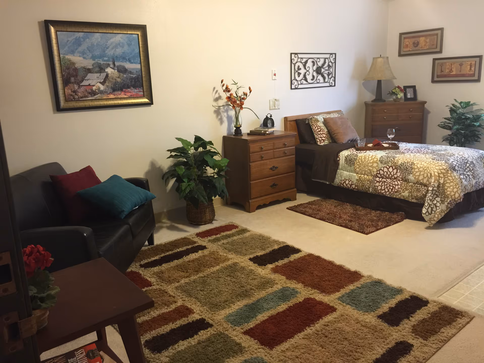 A furnished bedroom with a bed, dressers, an armchair, decorative rugs, plants, and wall art.